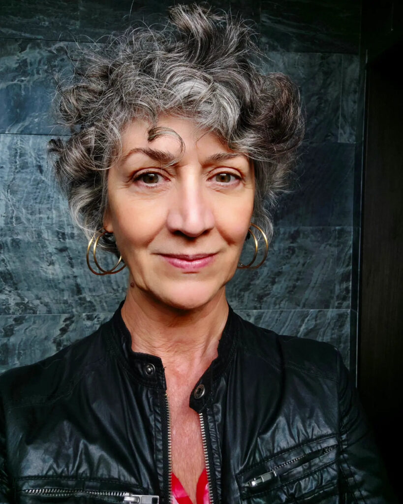 A woman with a short, messy pixie cut featuring curly, salt-and-pepper hair.