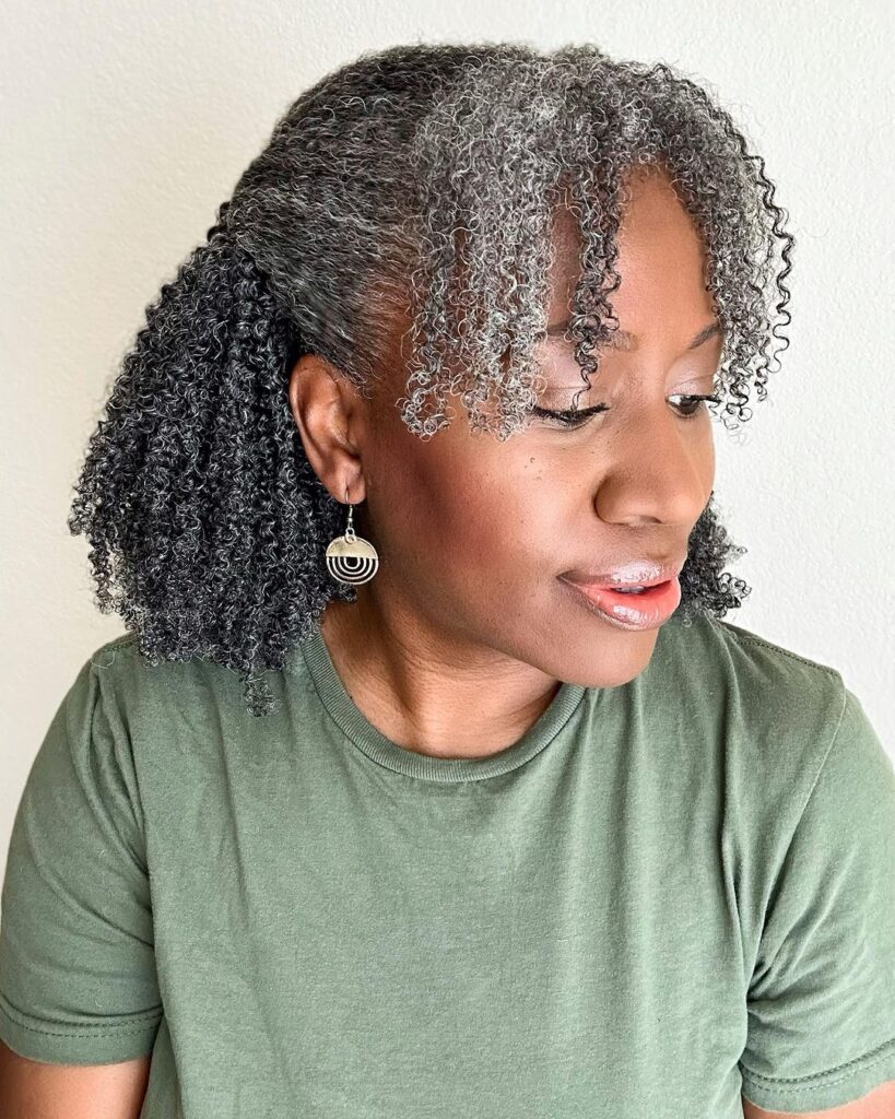 A woman with tight salt-and-pepper curls styled in a half-updo with curly bangs.