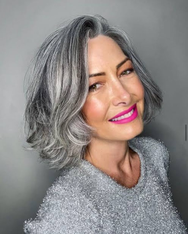 A smiling woman with a shoulder-length silver lob featuring voluminous, bouncy waves and a deep side part.