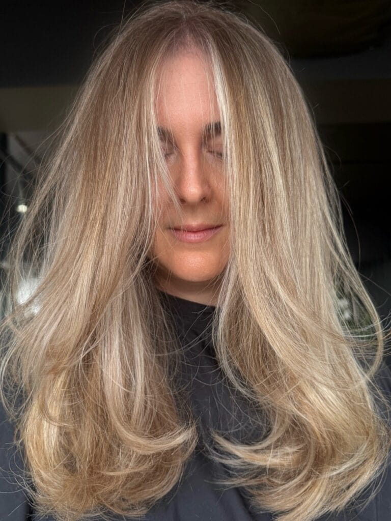 Long blonde layered hairstyle with voluminous body and softly curled ends.
