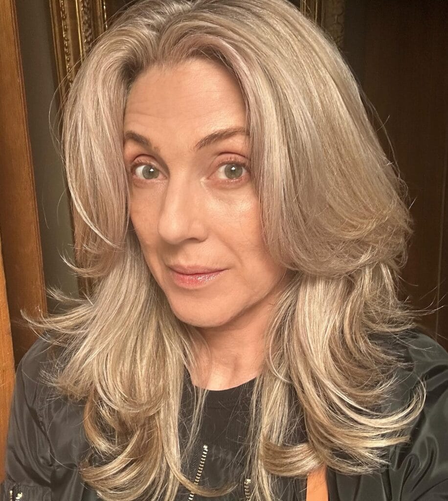 Medium to long blonde layered hair styled in a soft blowout with face framing volume.