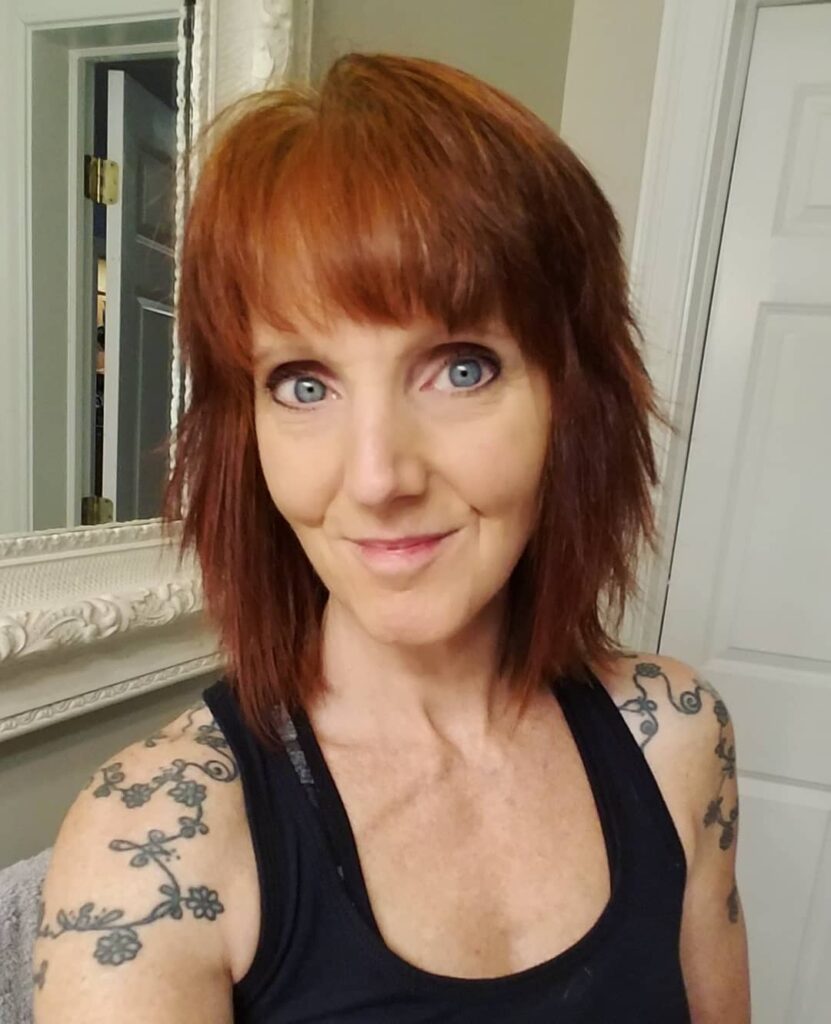 A woman with medium-length auburn hair styled in a layered shag with wispy bangs and a textured finish.