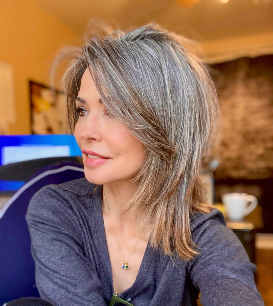 A woman with a medium-length layered hairstyle in salt and pepper tones featuring high volume and a side-swept fringe.