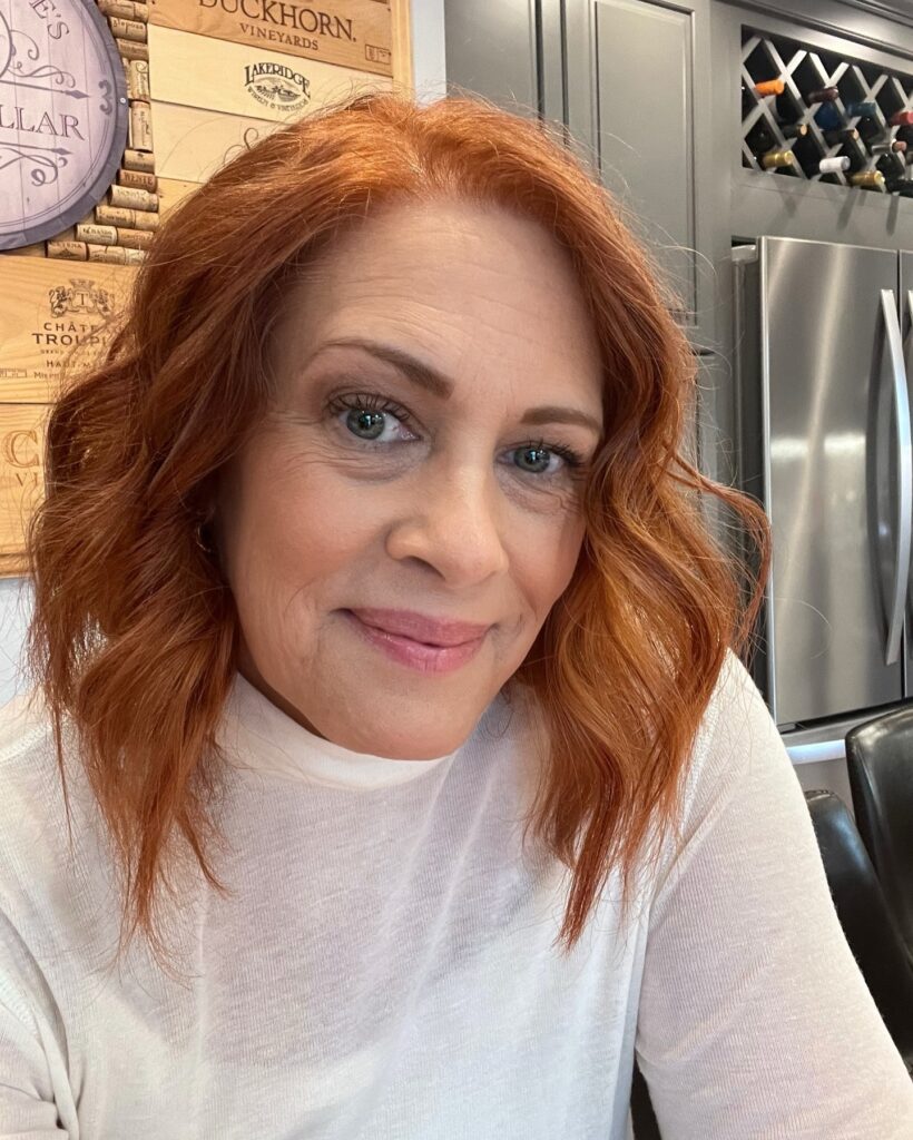 A woman with medium-length bright copper hair styled in soft, relaxed waves with a middle part.