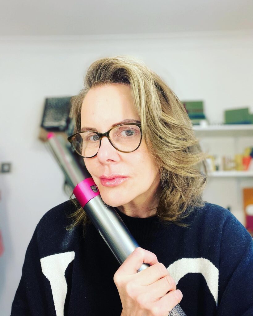 A woman with glasses holding a styling tool has medium-length ash-blonde hair with voluminous swept-back layers and a side part.