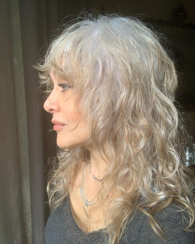 A side profile of a woman with medium-length wavy silver hair styled in a layered shag with wispy bangs.