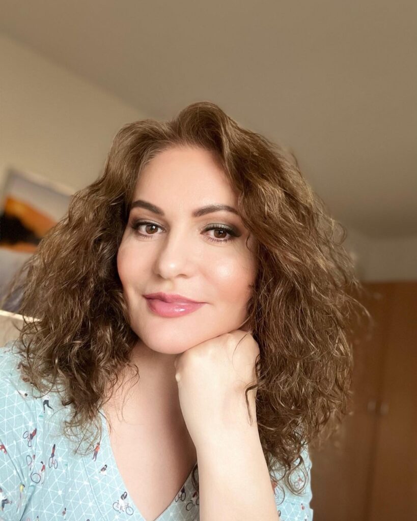 A woman with shoulder-length curly chestnut brown hair and a middle part.