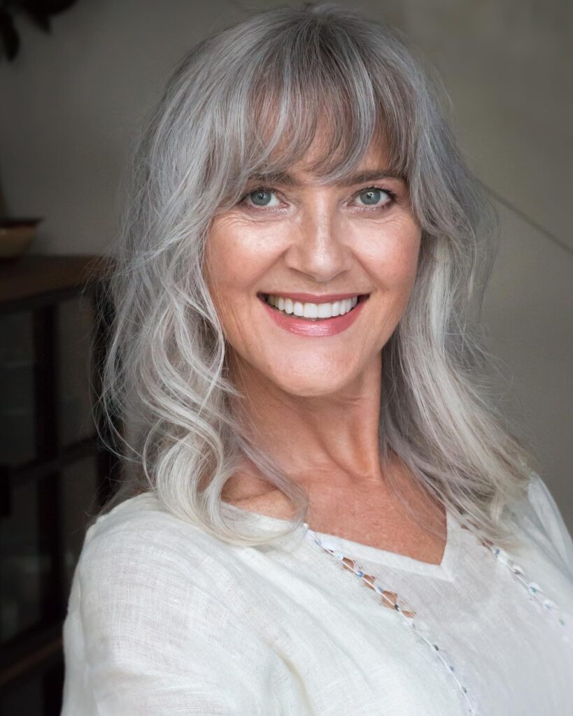 A smiling woman with shoulder-length silver hair styled in soft waves with light piecey bangs.