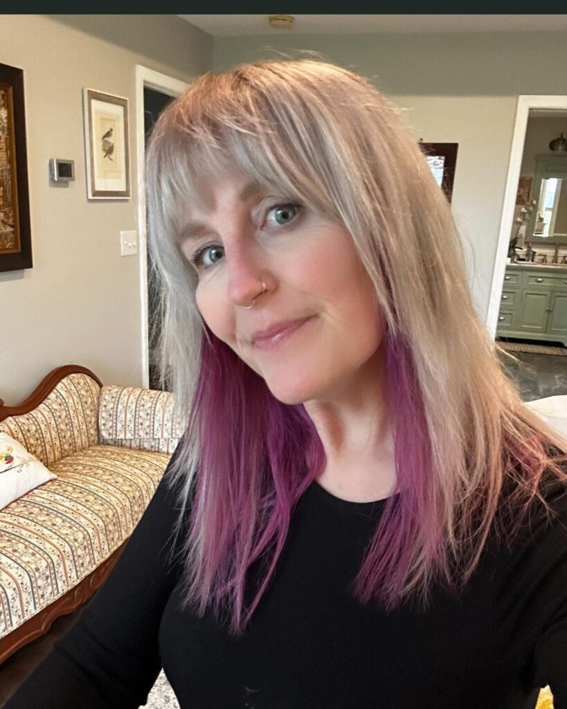 A woman with shoulder-length platinum blonde hair featuring purple-dyed underlayers and a full textured fringe.