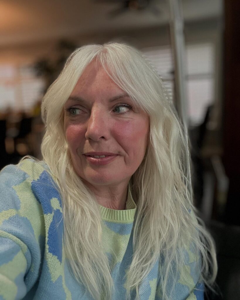 A woman with shoulder-length bright platinum hair styled in loose beachy waves with soft wispy curtain bangs.