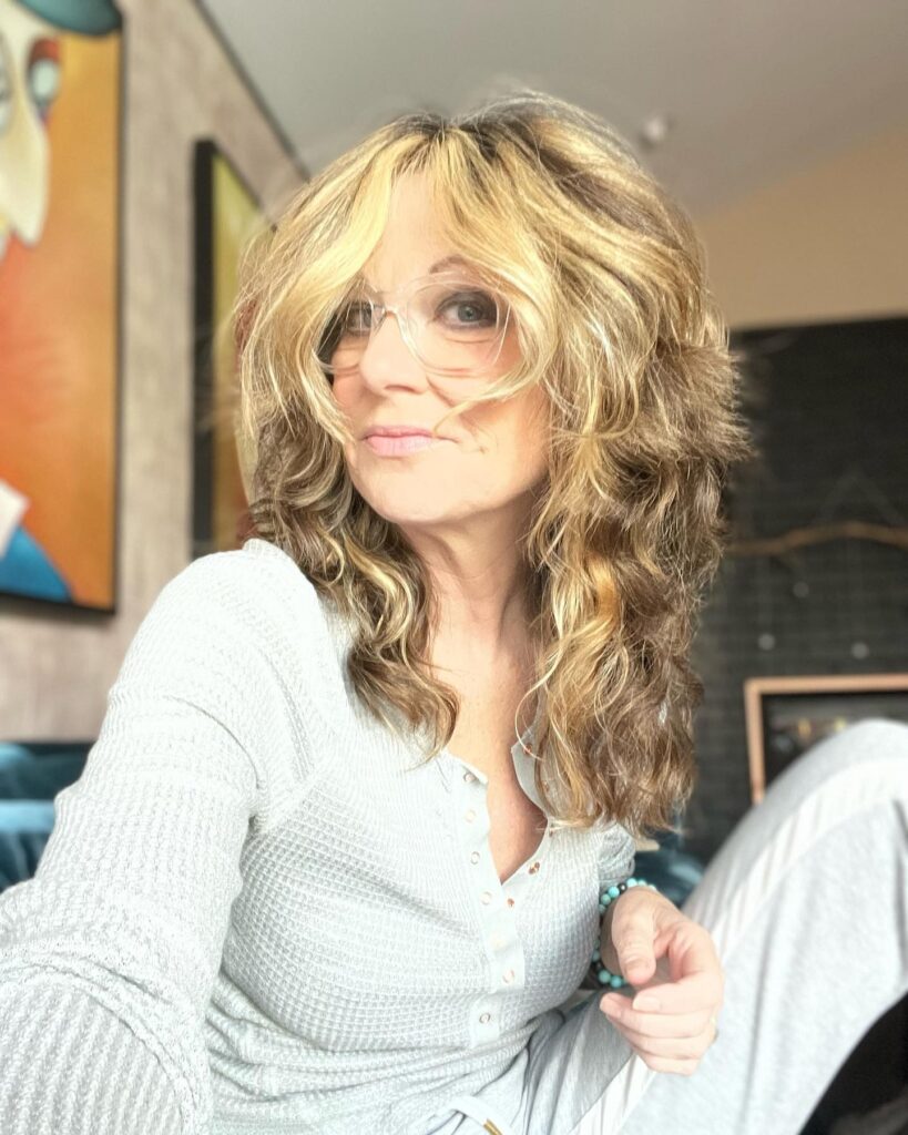 A woman with shoulder-length wavy honey-blonde hair in a layered shag cut with curtain bangs.