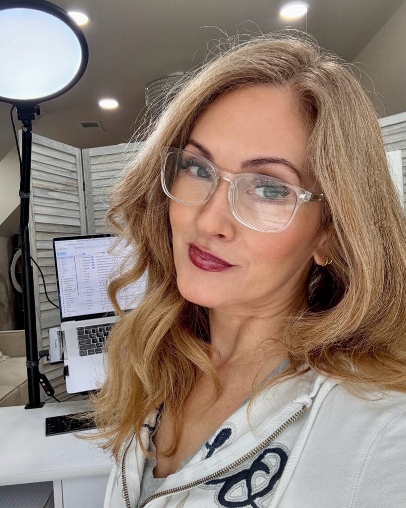 Shoulder length blonde hair with soft waves worn with clear glasses.