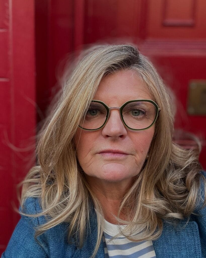 Shoulder length blonde wavy hairstyle worn with round glasses.