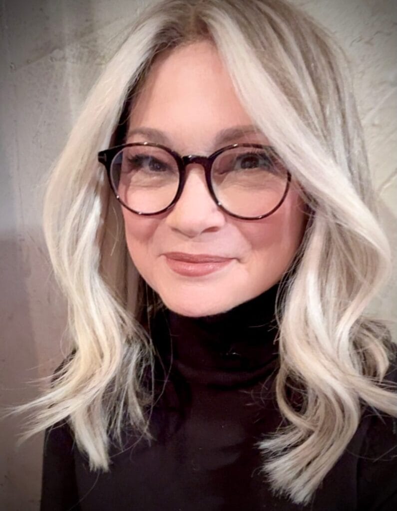 Shoulder length platinum blonde hair with loose waves worn with round glasses.