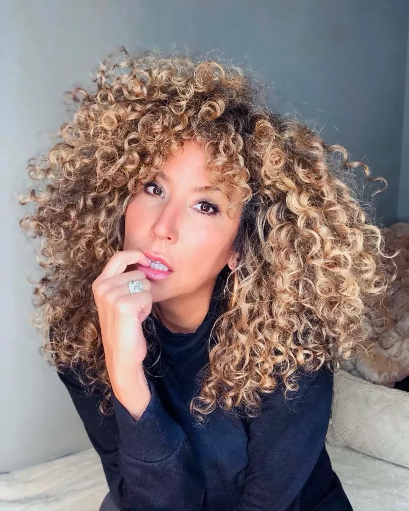 Tight curly layered hairstyle with voluminous shape and golden blonde tones.