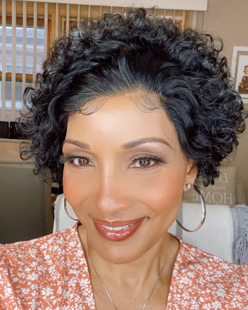 A smiling woman with a short, jet black, curly hairstyle featuring high volume and soft wisps at the hairline.