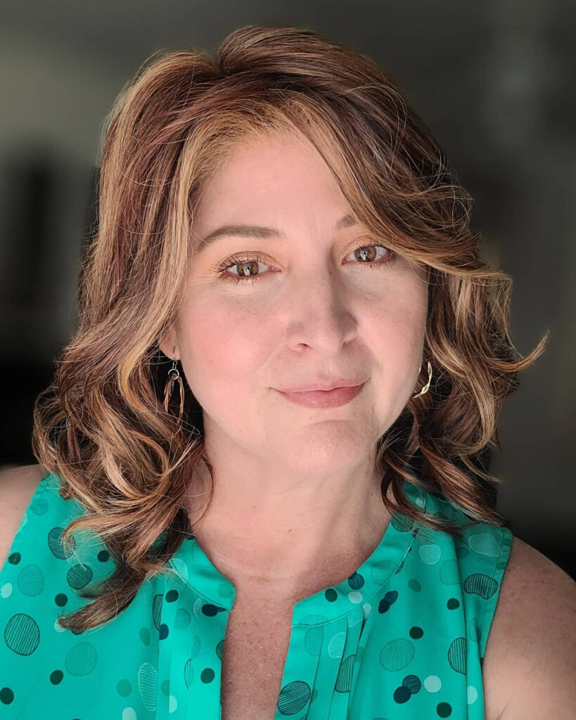 A woman with medium-length wavy auburn hair featuring golden highlights and a side-swept fringe.