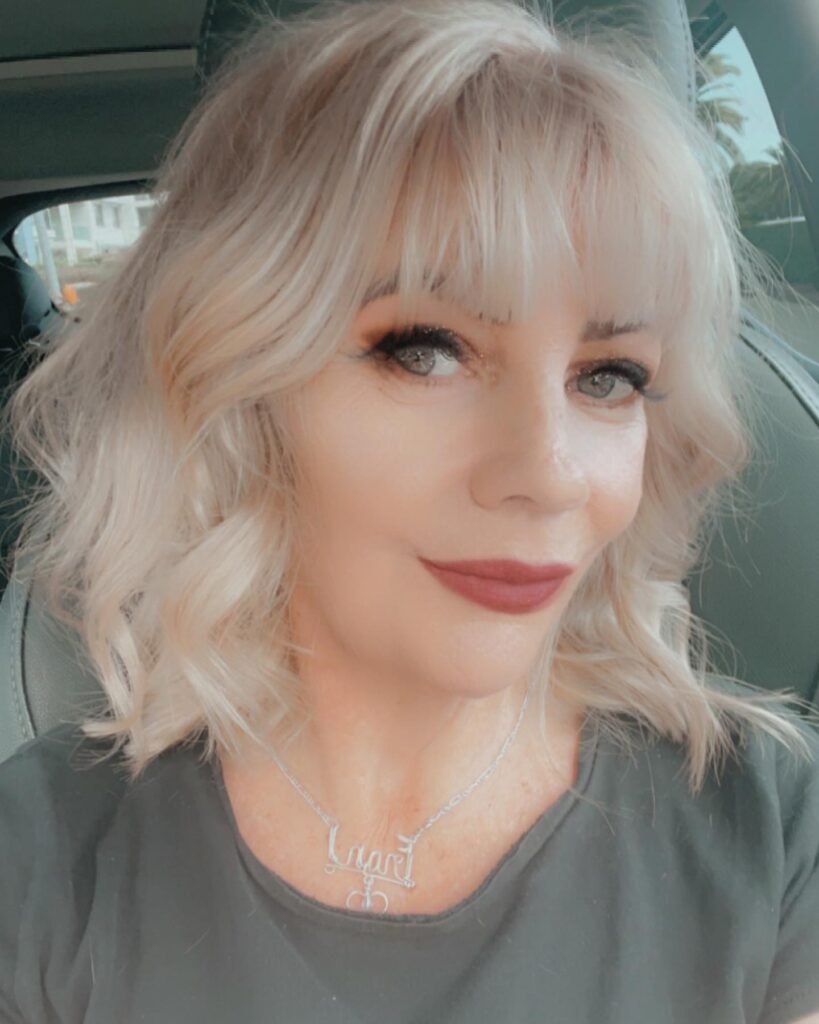 A woman with a shoulder-length creamy blonde shaggy haircut featuring a full wispy fringe and textured waves.