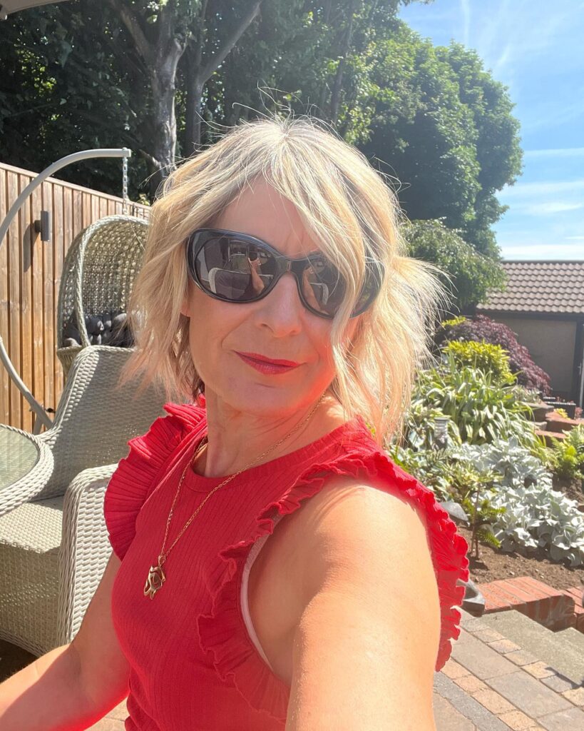 A woman outdoors wearing sunglasses and a red top with a tousled, sandy blonde chin-length bob and wavy texture.