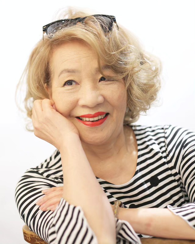 A smiling woman with voluminous honey blonde curls and a deep side part wearing a striped shirt.