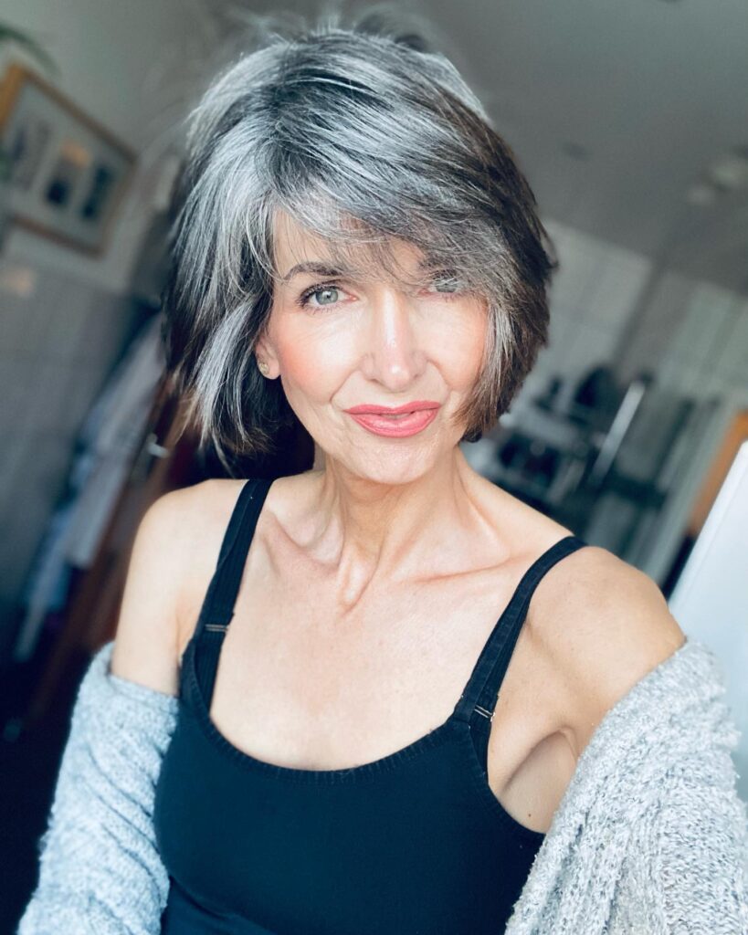 A woman with a textured salt-and-pepper chin-length bob and a long side-swept fringe wearing a black tank top and a gray cardigan.