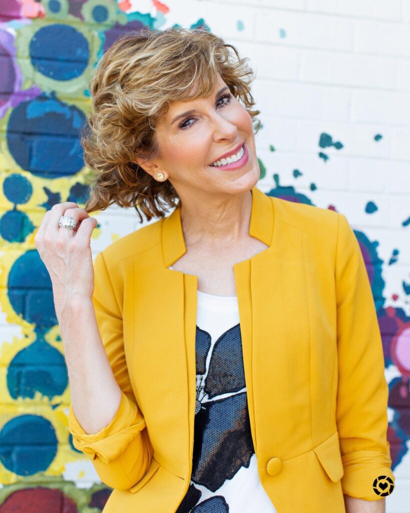 A smiling woman with a layered golden brown curly hairstyle wearing a vibrant yellow blazer against a colorful mural background.