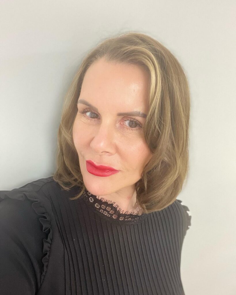 A woman with a sleek ash brown bob and subtle golden highlights wearing a black ruffled top and bold red lipstick.