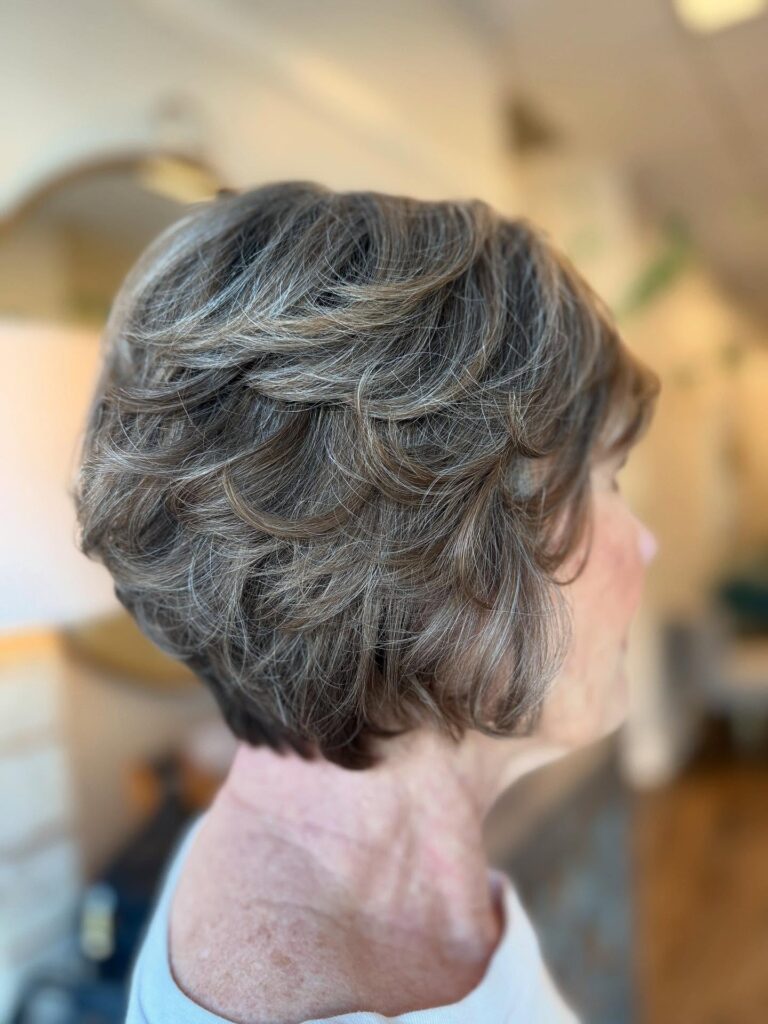 A side profile of a woman with a short feathered brunette bob featuring silver highlights and voluminous swept-back layers.