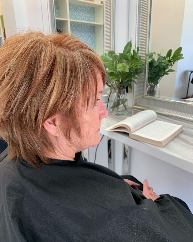 A side view of a woman with a choppy, shaggy copper hairstyle featuring golden highlights and wispy bangs.