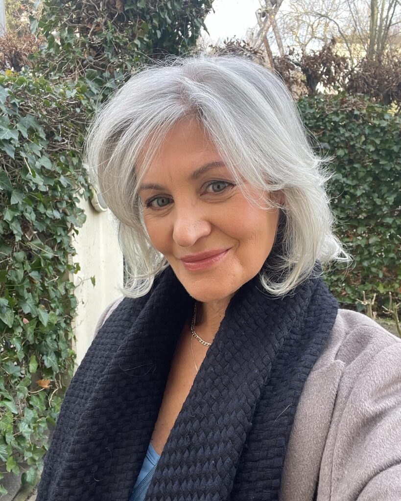 A smiling woman with a feathered silver lob hairstyle featuring flicked ends and a side sweep wearing a black scarf and gray coat.