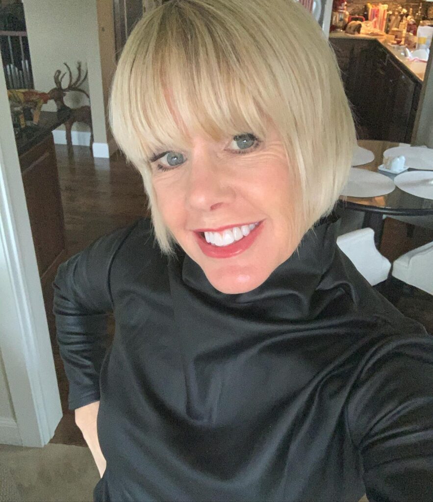 A smiling woman with a sleek, sandy blonde, chin-length bob and full, straight bangs, wearing a black turtleneck.