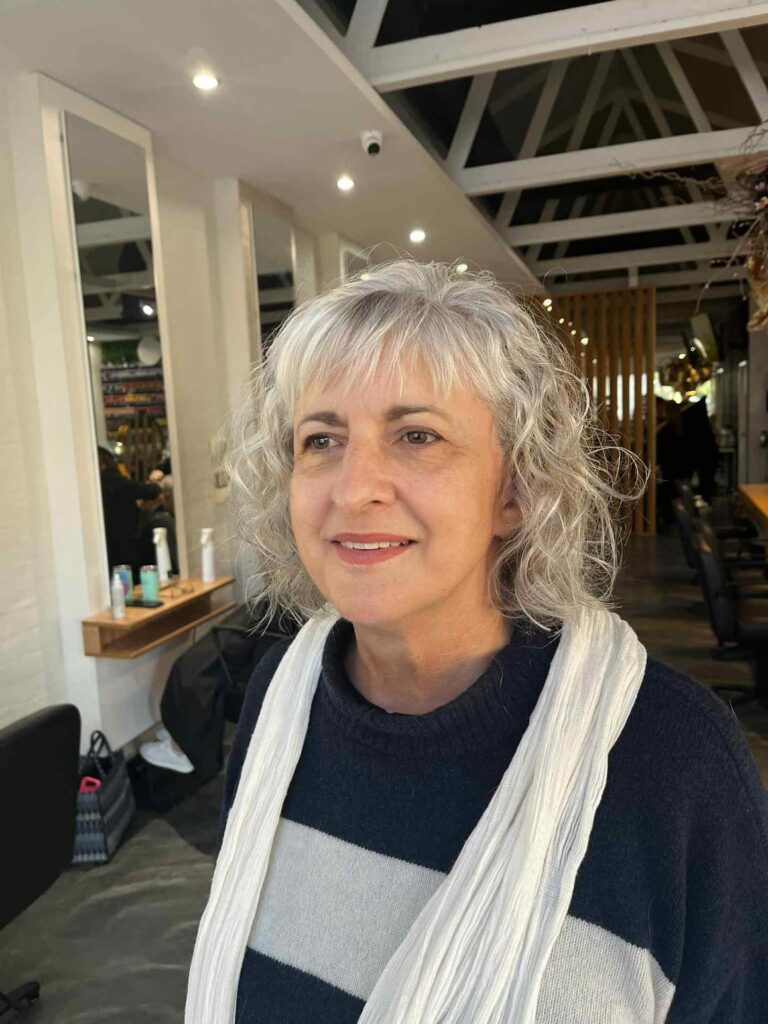 A woman with shoulder-length wavy silver hair featuring a light wispy fringe, smiling in a salon.