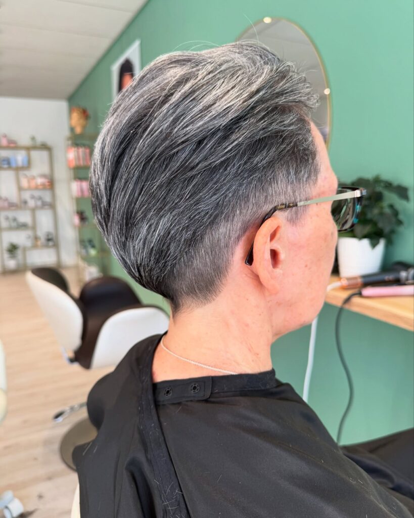 A close-up side view of a woman with a tapered silver and black pixie cut featuring longer layers swept back at the crown.
