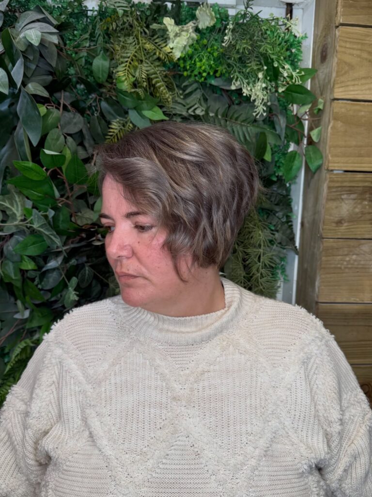 A woman with an asymmetrical wavy bob in an ashy mushroom brown shade, looking to the side against a leafy background.