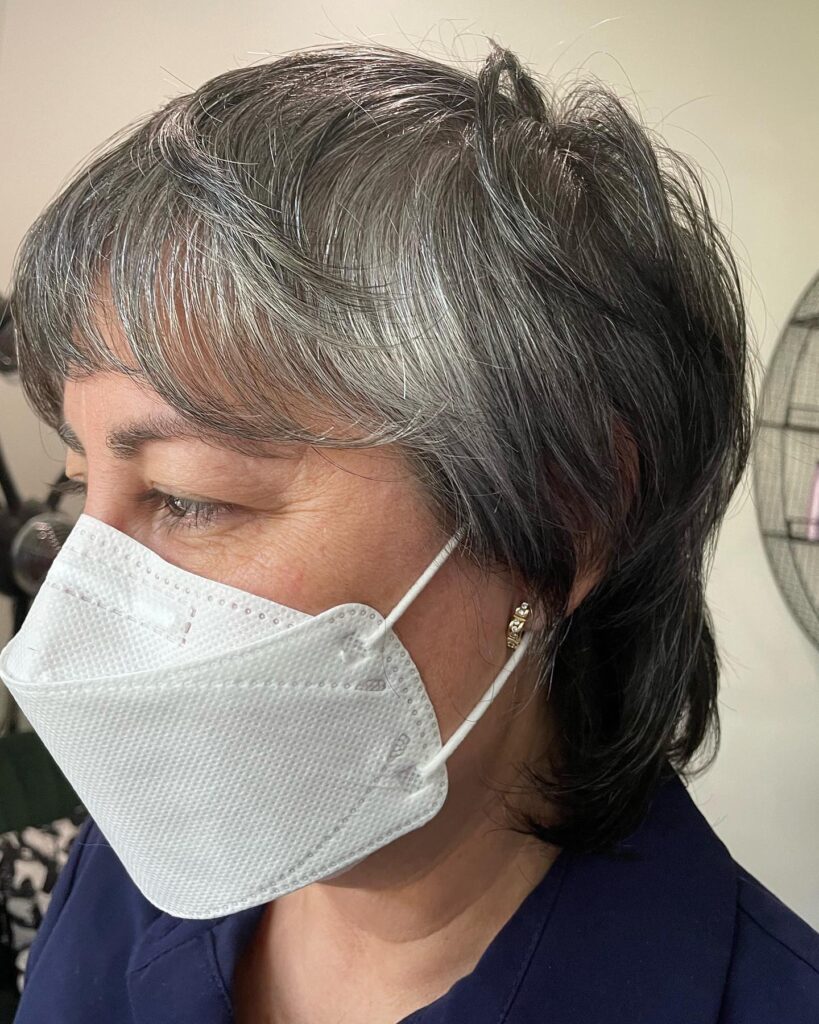 A close-up side view of a woman wearing a white mask with a layered charcoal and silver shaggy hairstyle and wispy bangs.