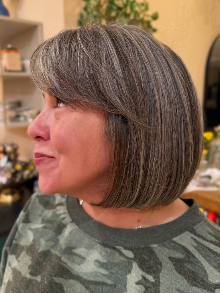 A side profile of a woman with a rounded chin-length bob in a salt-and-pepper grey shade featuring a side-swept fringe.