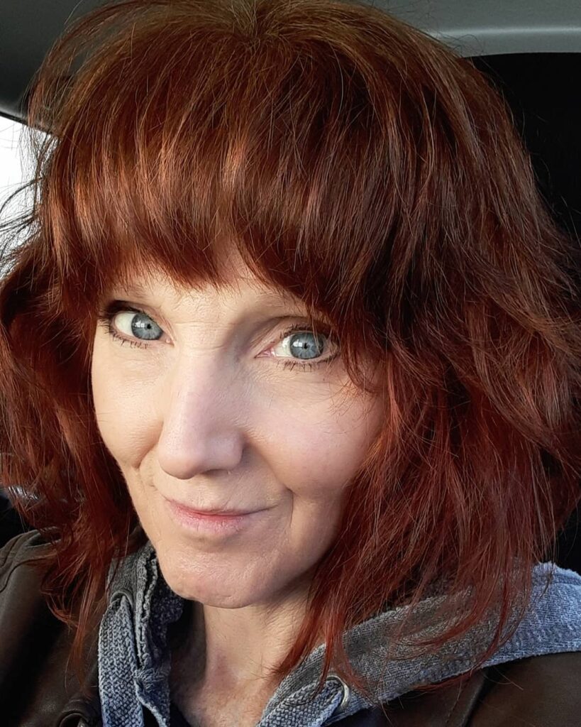 A middle-aged woman with vibrant red hair styled into a shoulder-length shaggy bob with blunt bangs.