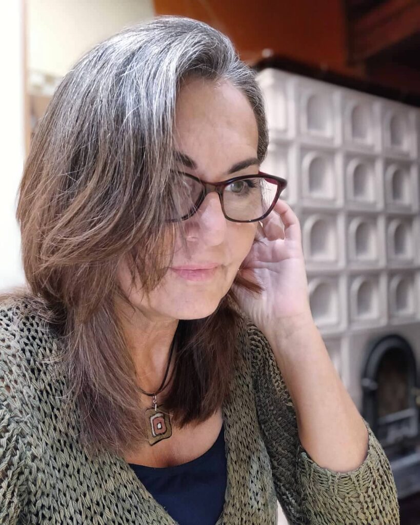 A middle-aged woman with glasses wearing natural silver and brown hair in a medium-length layered bob with long side-swept bangs.