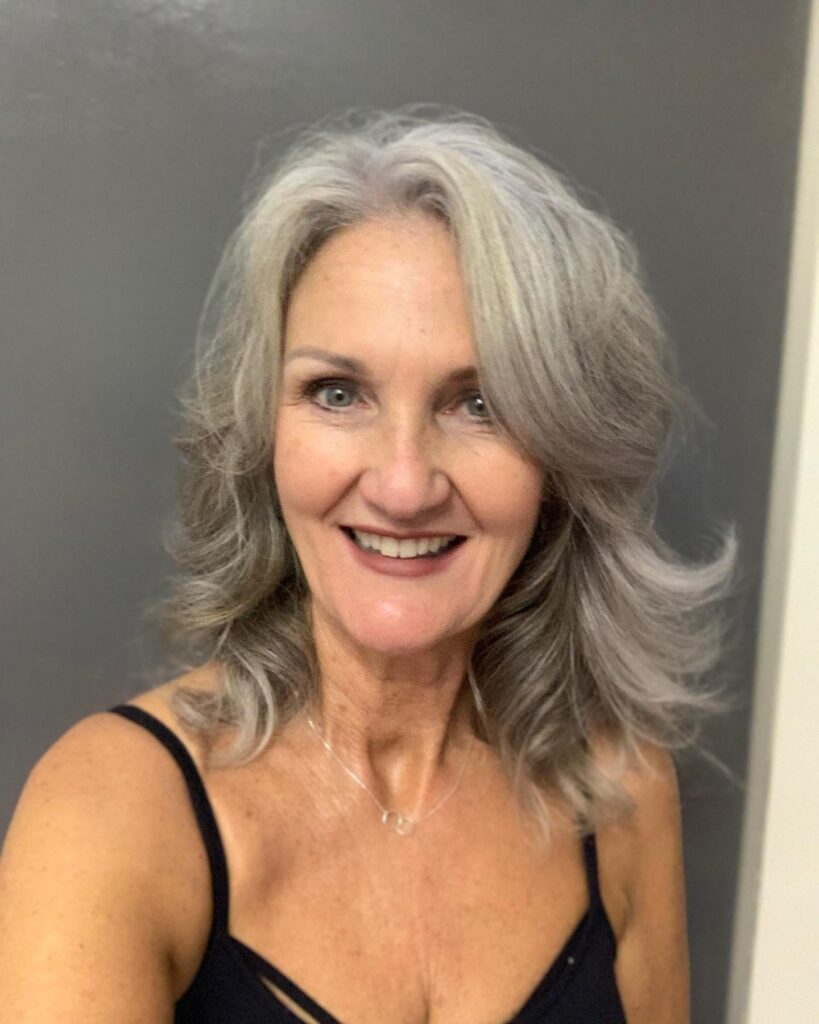 A smiling woman with shoulder-length layered silver hair and voluminous side-swept bangs.