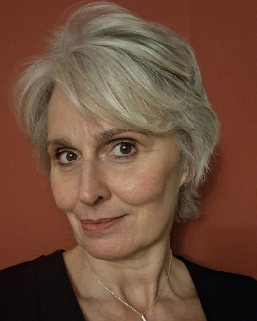 A woman with a soft silver pixie cut and feathered side-swept bangs posing against a reddish background.