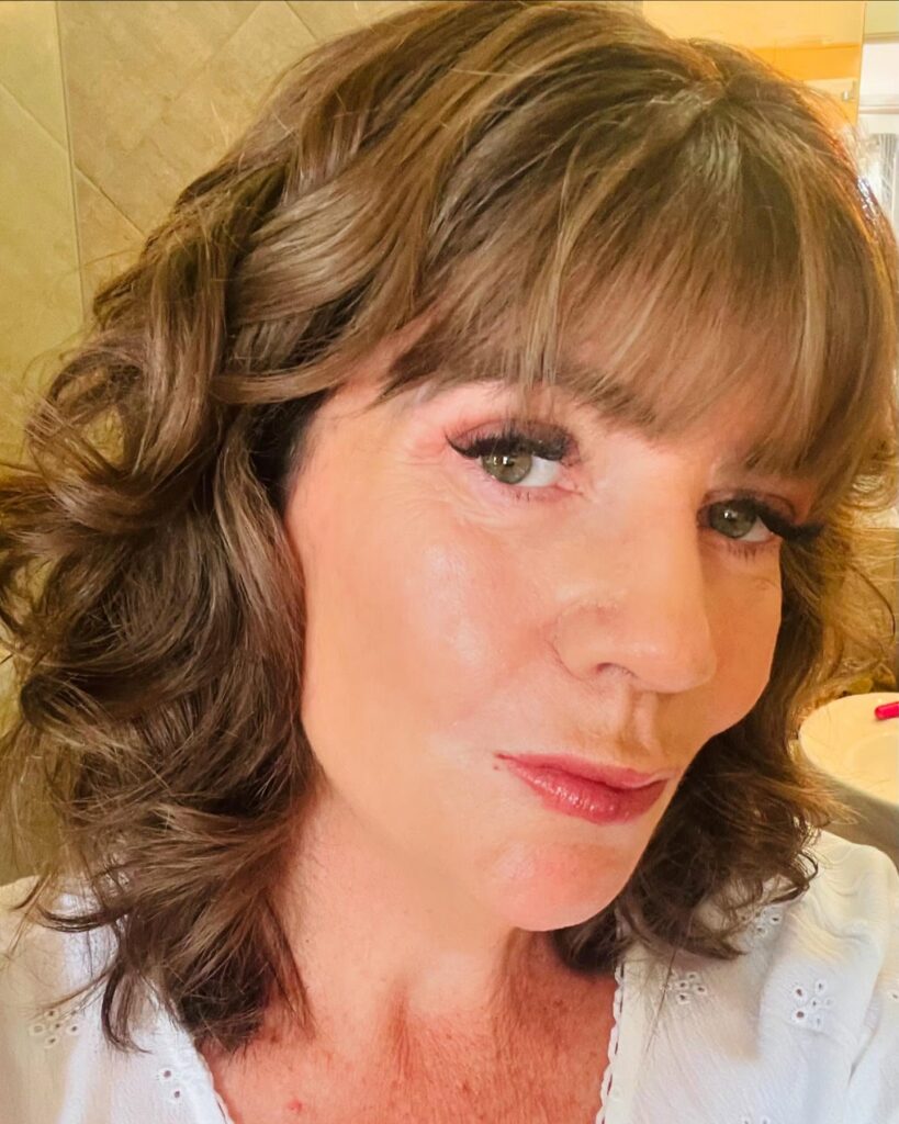 A woman with shoulder-length curly brown hair and full bangs looking directly at the camera.
