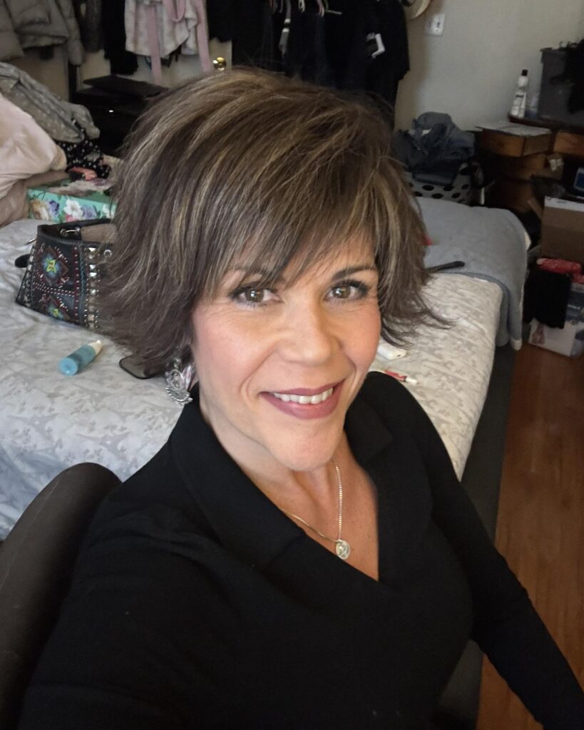 A smiling woman with a short, layered ash brown bixie cut and choppy jagged bangs.
