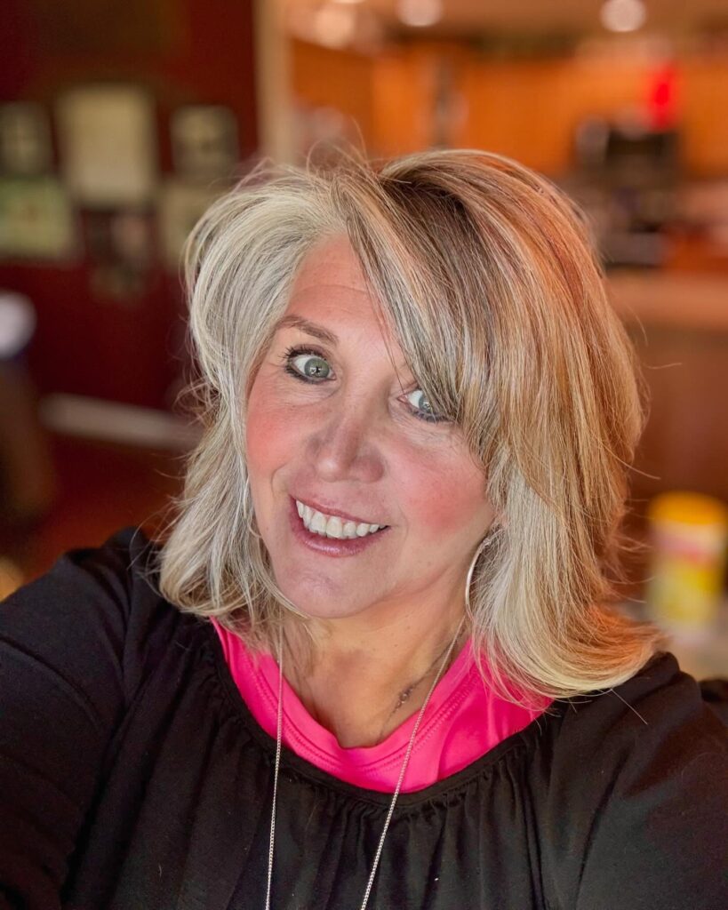 A smiling woman with multi-tonal ash-blonde shoulder-length hair and a deep side-swept fringe.