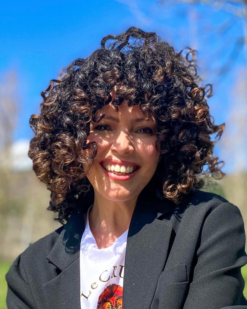 A smiling woman with voluminous shoulder-length dark curly hair and curly bangs outdoors in the sun.