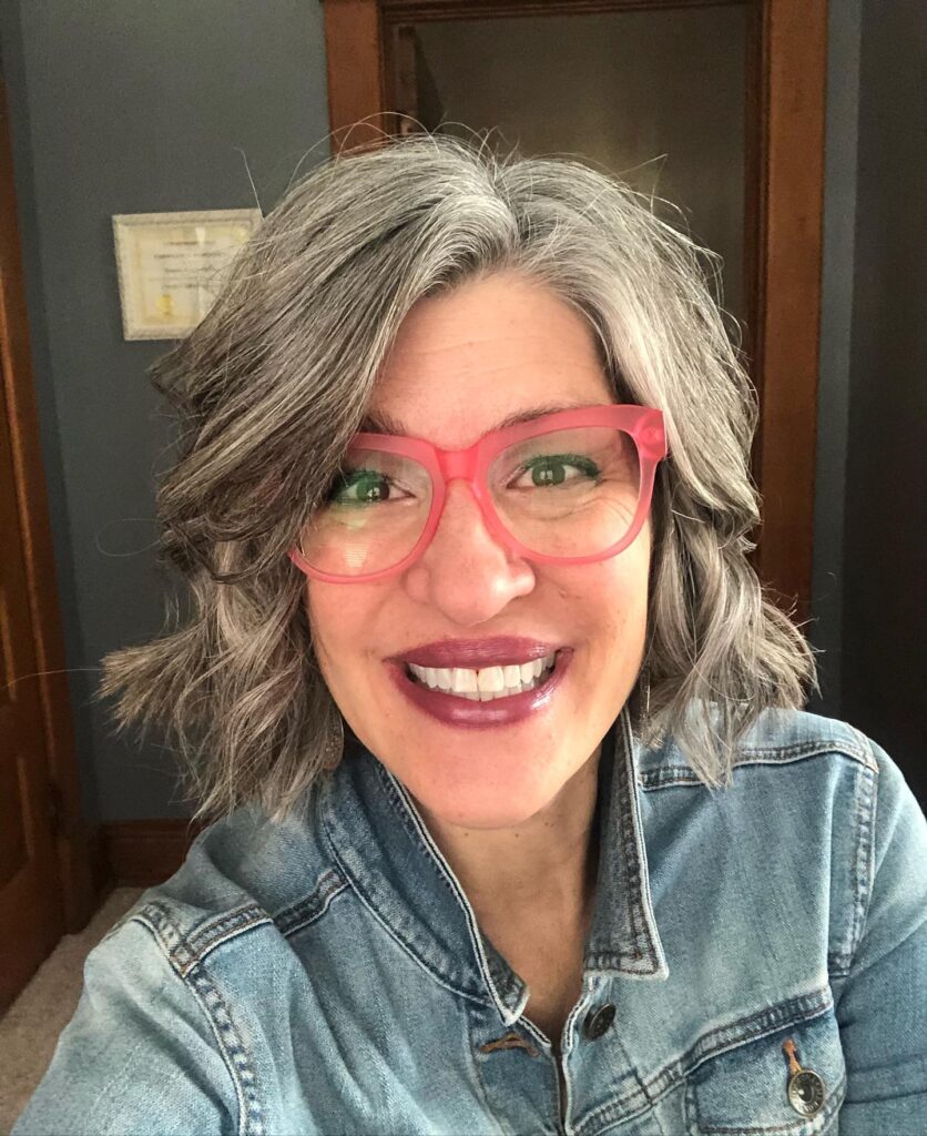 A smiling woman with a wavy salt-and-pepper bob and side-swept bangs wearing bright pink glasses.