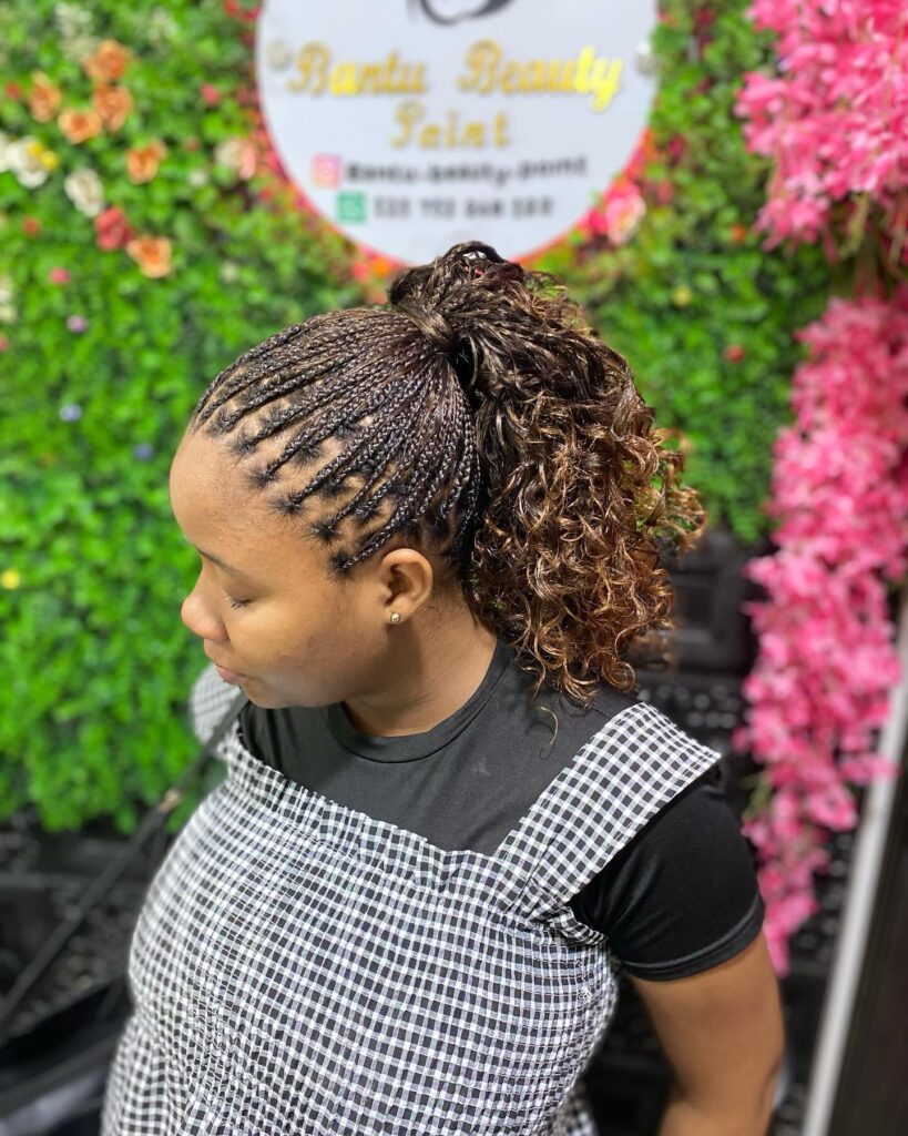 A woman wearing a high ponytail featuring small braids and voluminous brown curls with golden highlights.