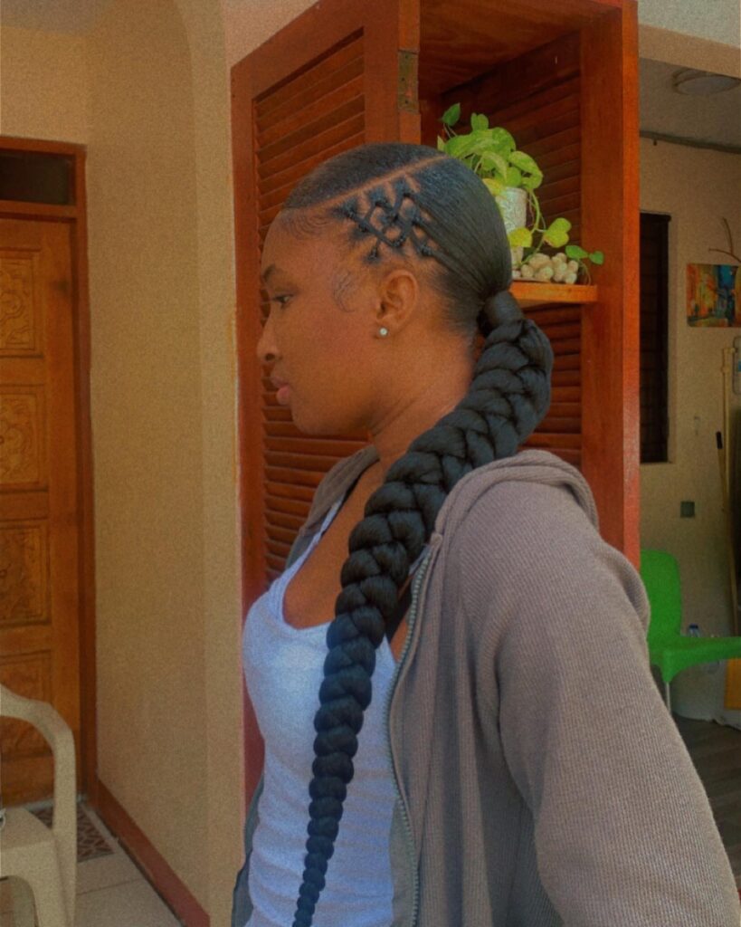 A woman in profile wearing a very long and thick black braid styled as a low ponytail with a decorative criss-cross pattern on the side of her head.
