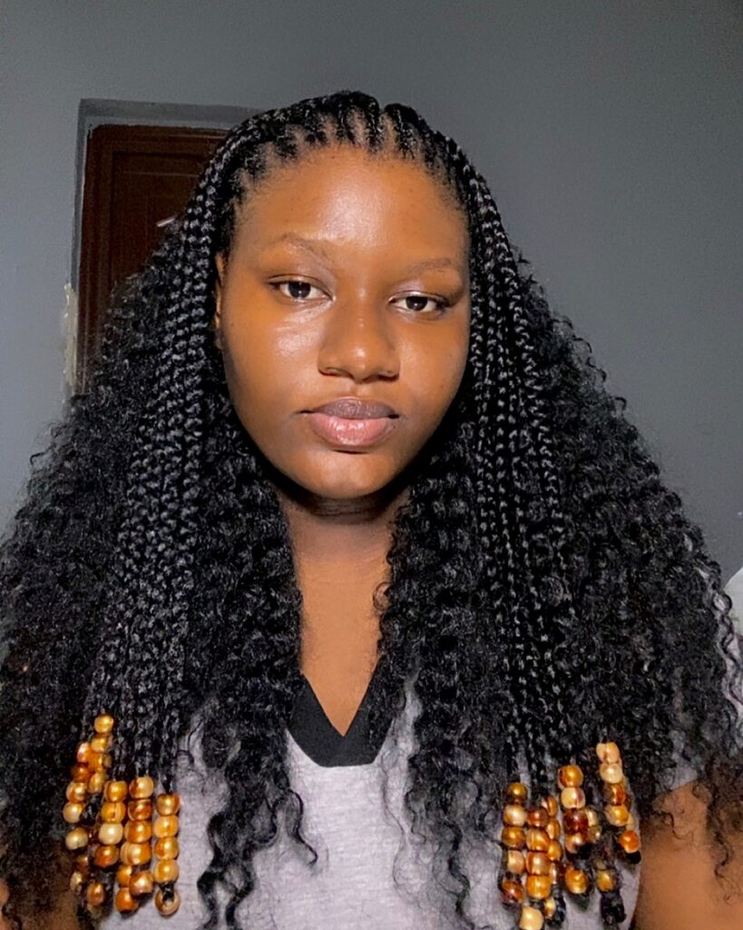 A woman with long black micro braids and twists, featuring stacked wooden beads at the ends of the front sections.