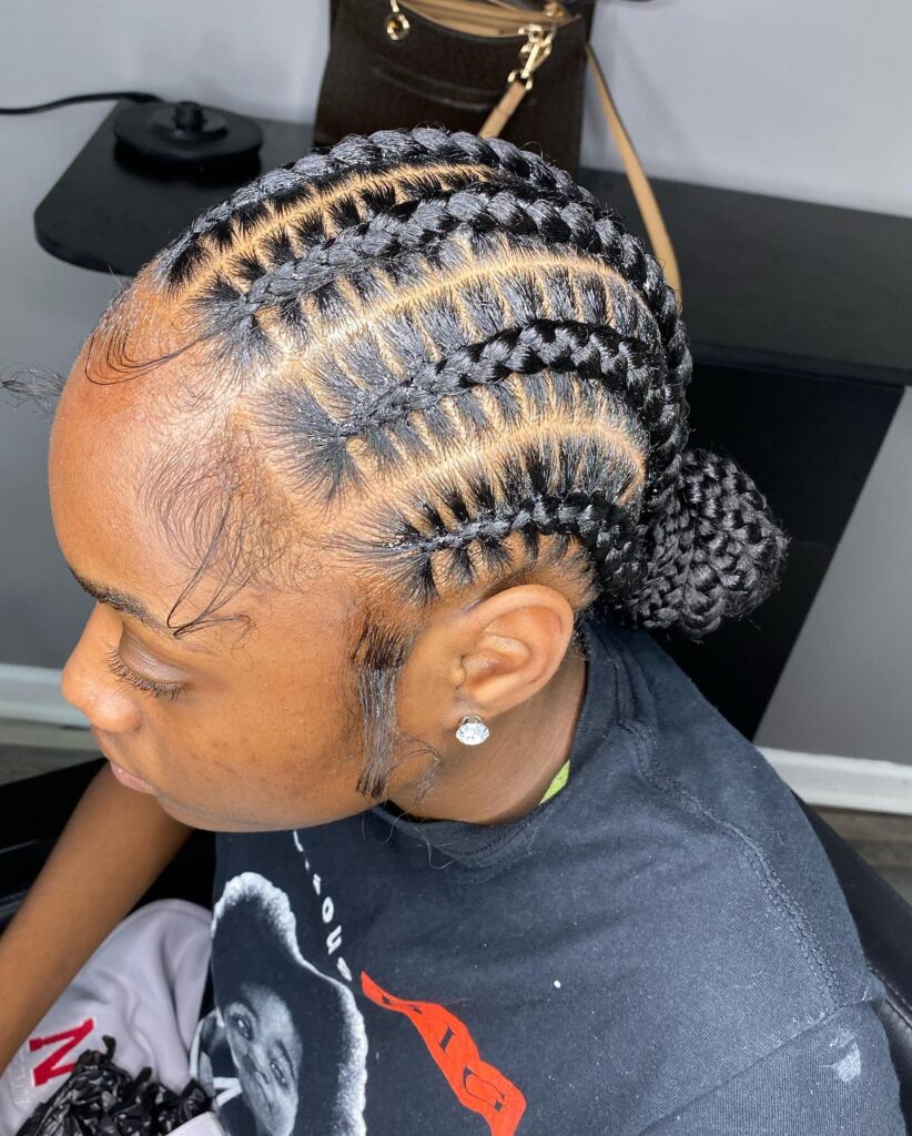 A side view of a woman with neat, stitched braids curving into a low braided bun with styled edges and a single curl on her cheek.