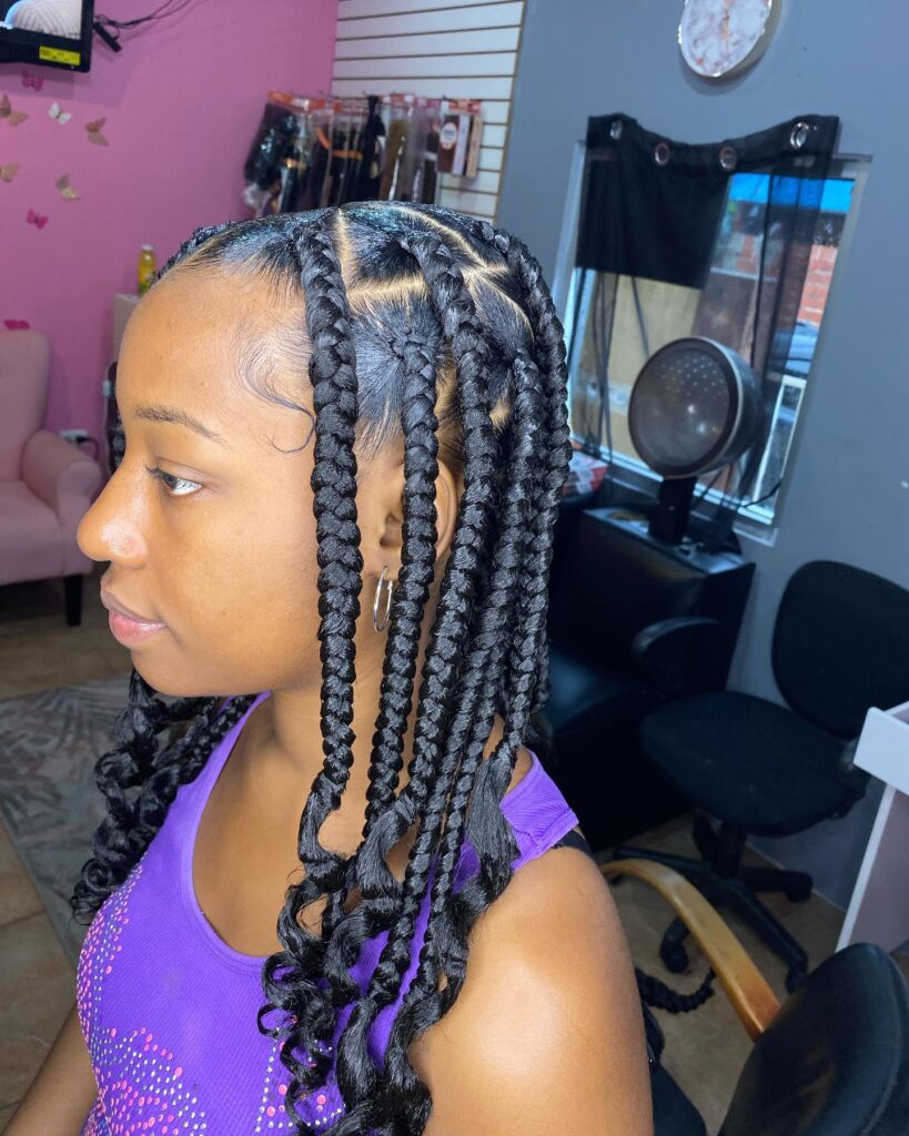 A young woman with thick black knotless braids featuring large square parts and curly spiraled ends.
